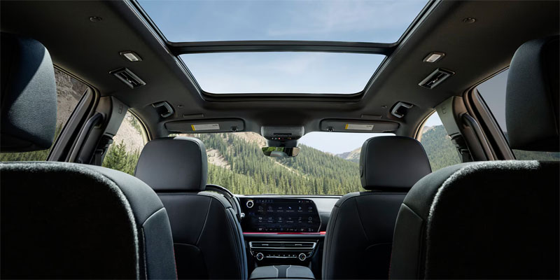 Interior view of a 2026 Chevy Traverse with a panoramic sunroof, featuring front seats and dashboard, overlooking a scenic mountainous landscape through the glass.