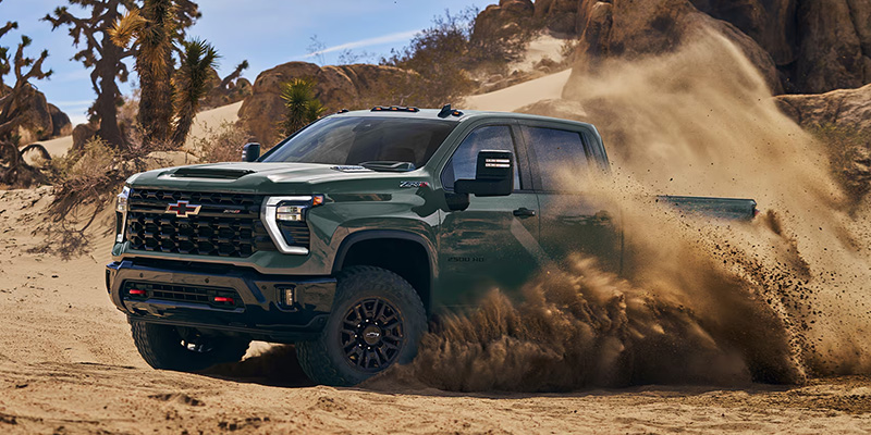Green Chevrolet Silverado HD pickup truck driving through sandy desert terrain, kicking up dust while showcasing its rugged off-road capability and durable design.