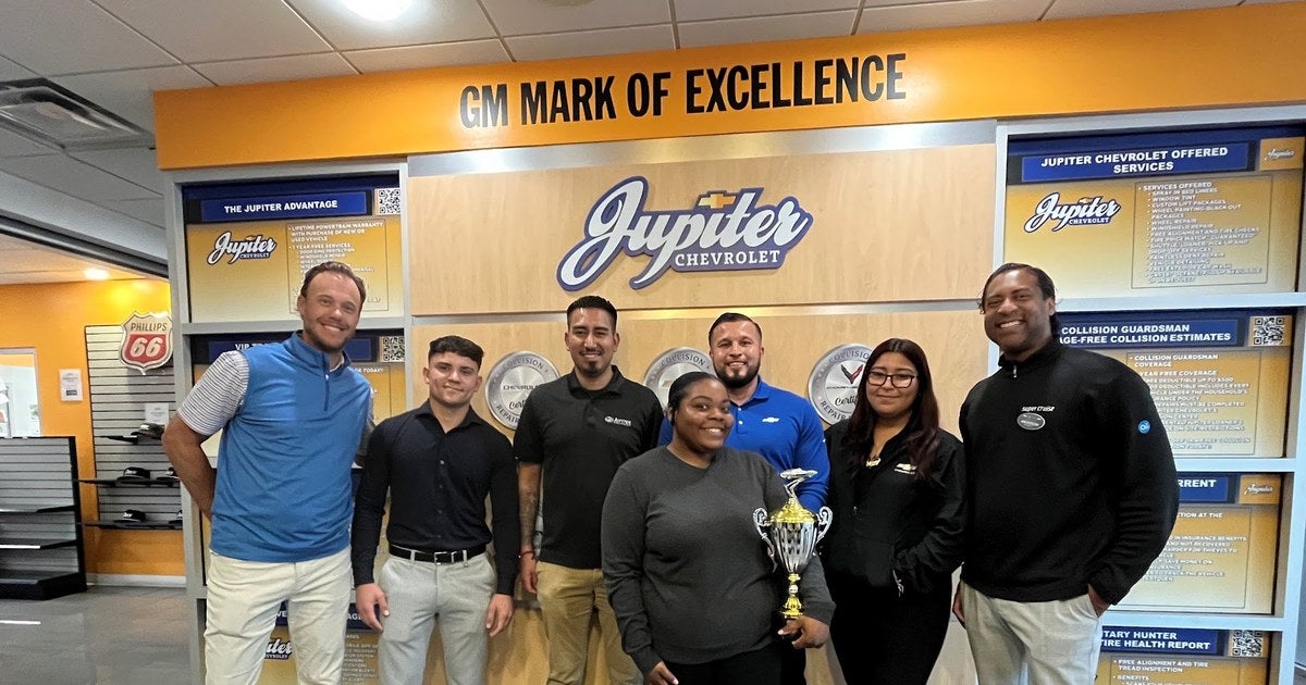 The Jupiter Chevrolet team in the Garland, TX showroom beneath the GM Mark of Excellence banner